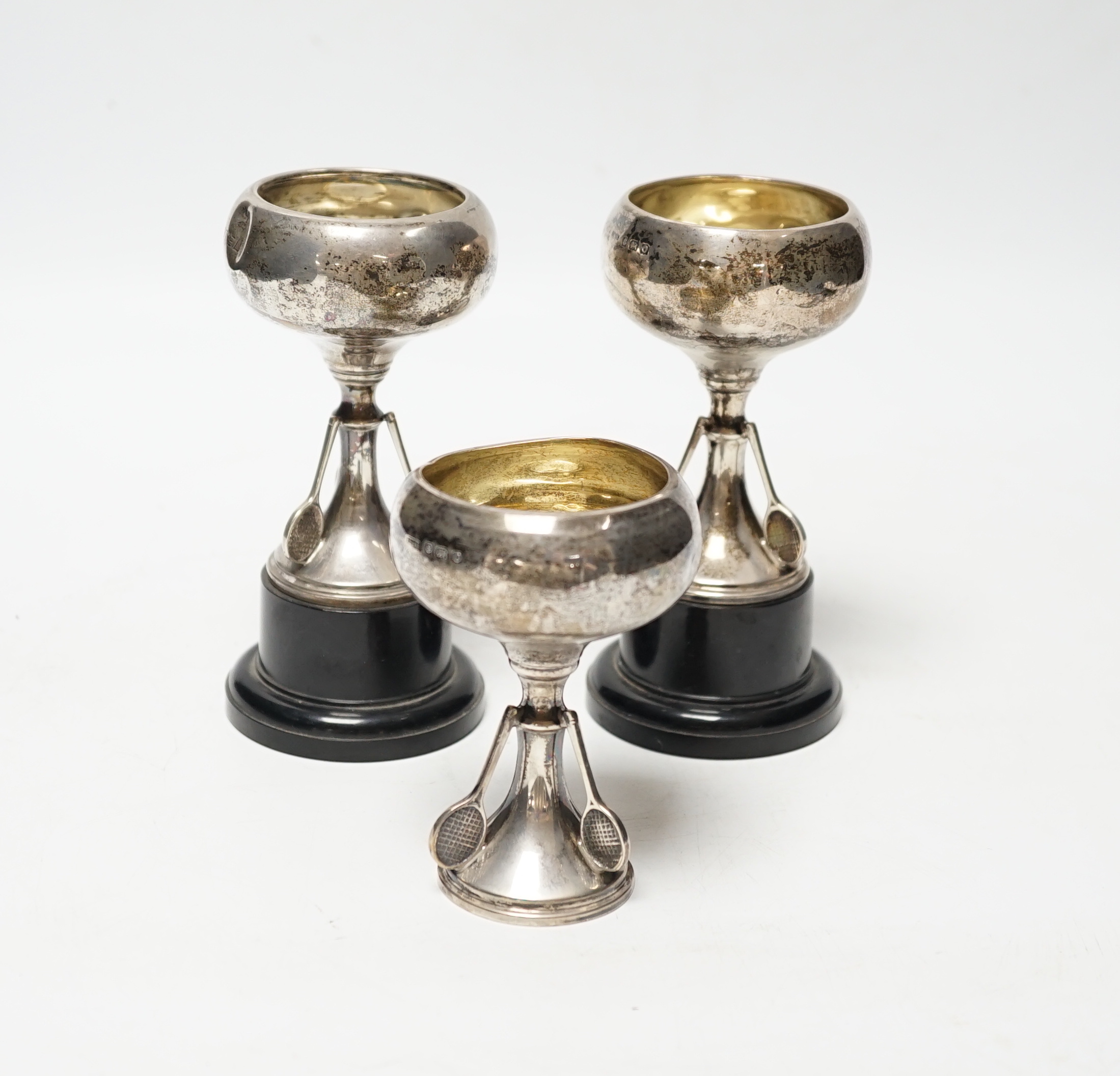 A set of three small George V silver tennis related trophy cups, with racket supports and engraved inscriptions, E & J Leek, 92mm, two with plinth bases.                                                                   