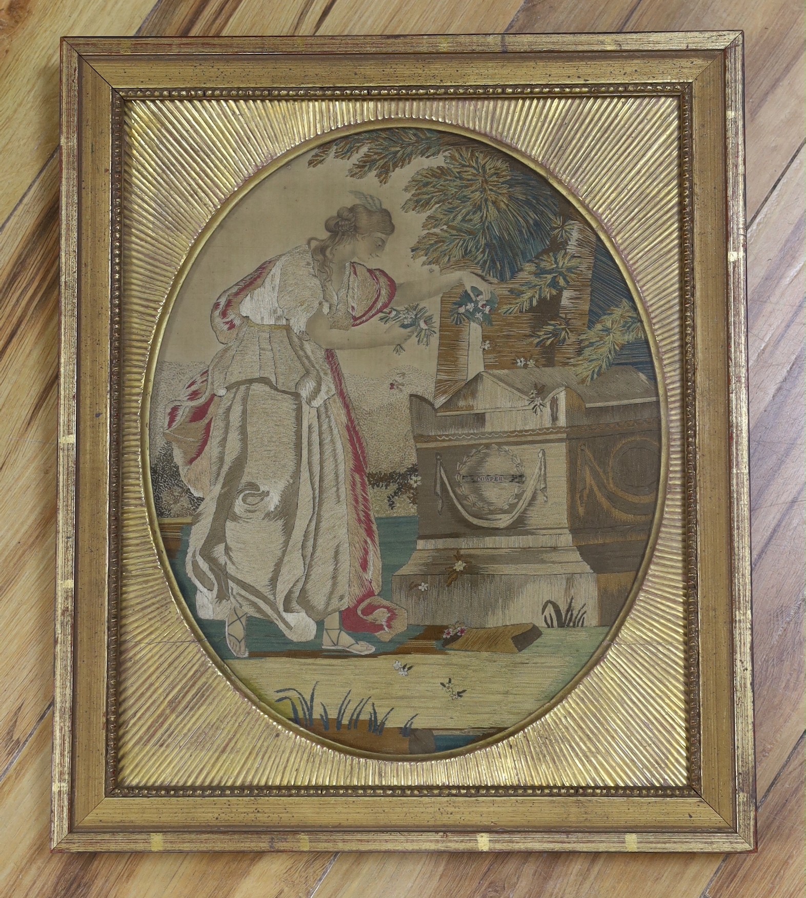 A gilt framed Regency oval silkwork picture of a classical maiden scattering flowers at a tomb, labelled Cowper, probably commemorating the death of the poet William Cowper in 1800, 30x25cm                               