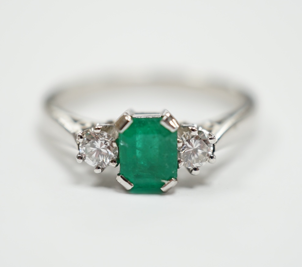 A modern 18ct white metal, single stone emerald and two stone diamond set ring, size N, gross weight 4 gram.                                                                                                                