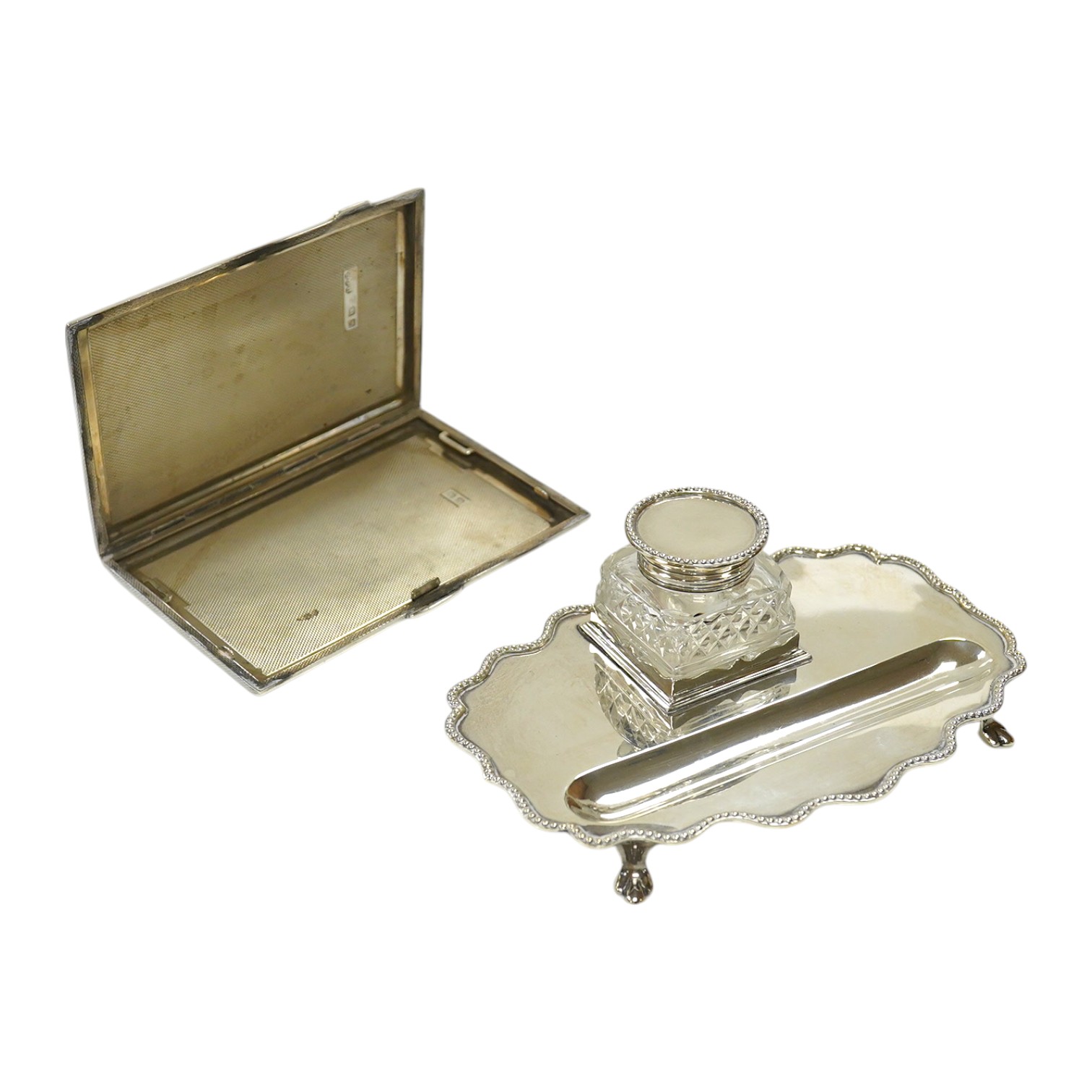 An Edwardian silver shaped oval inkstand, with wavy border, silver mounted glass well and pen recess, Skinner & Co, London, 1908, 15.8cm, together with a later engine turned silver cigarette case, 10.6oz. Condition - fai