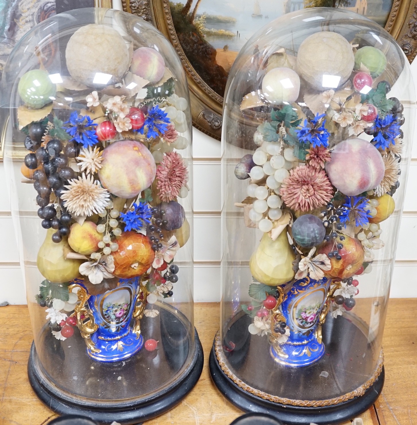 A pair of Victorian displays of painted papier mache fruit in vases, under glass domes, 58cms high                                                                                                                          