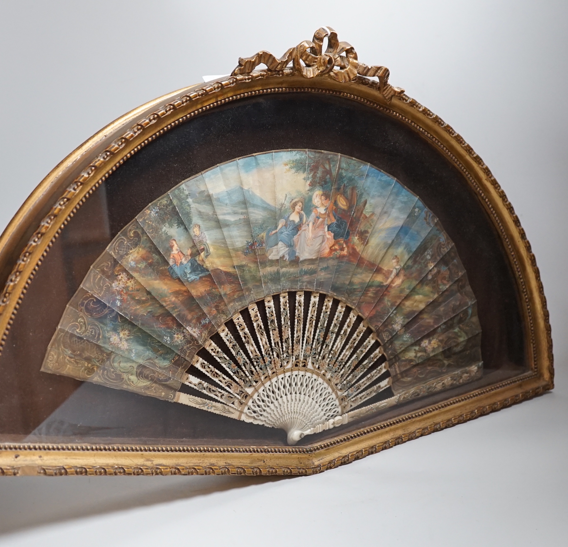 A 19th century French bone fan with pierced decoration, hand painted with figures wearing 18th century dress before a landscape, framed, the frame 62cm wide                                                                