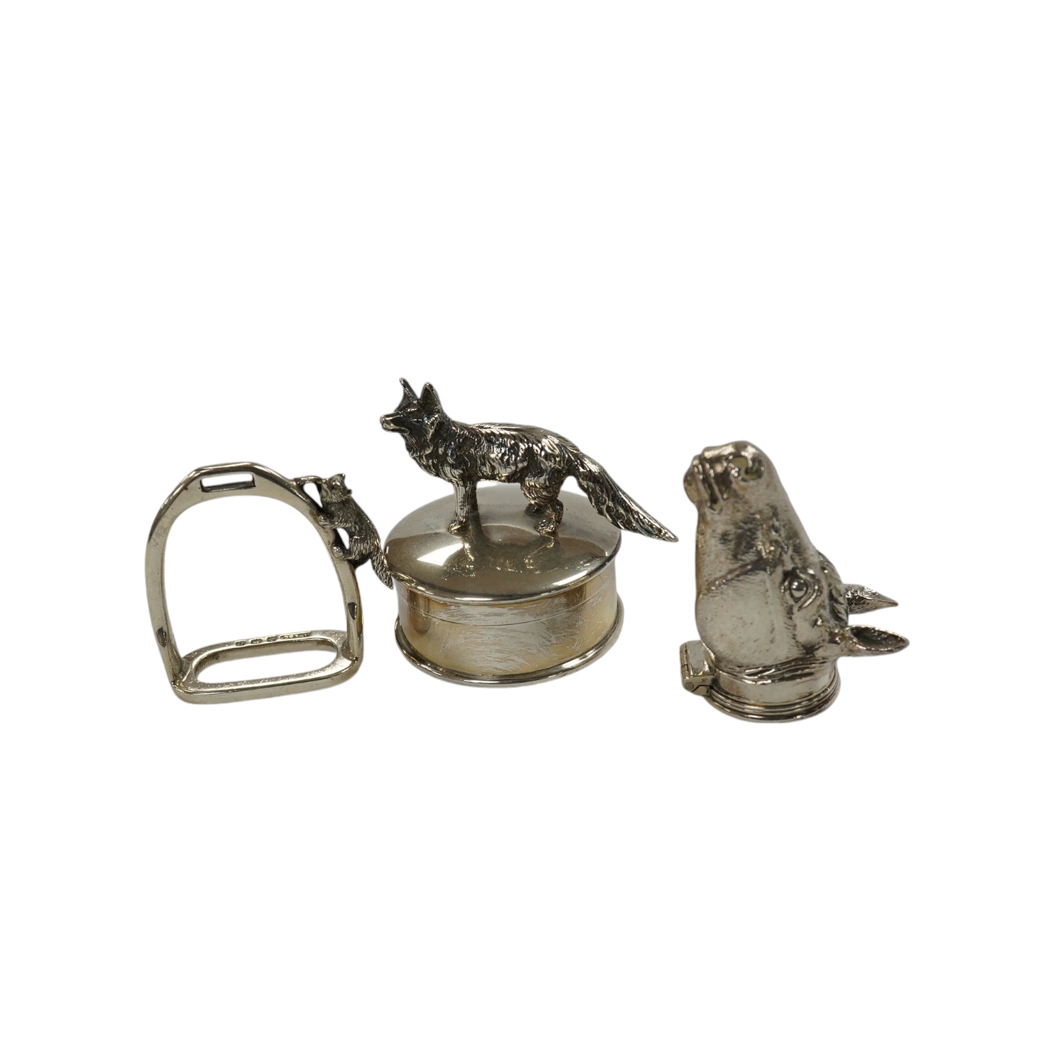 A George VI silver napkin ring modelled as a stirrup, surmounted with a fox, Birmingham, 1937, 46mm, together with a later silver horses head snuff box and a silver pill box with fox surmount. Condition - fair to good   