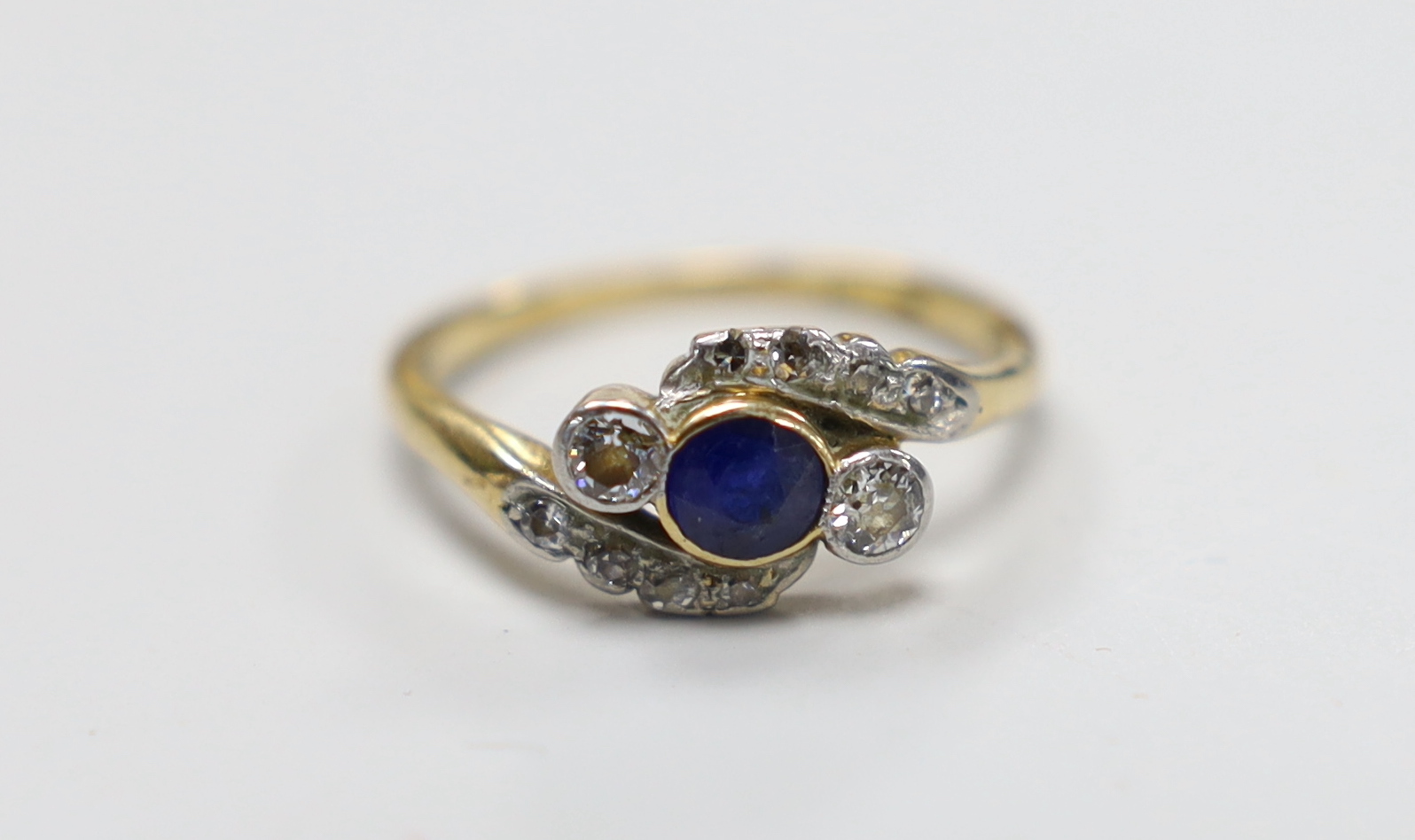 An 18ct, single stone sapphire and two stone diamond crossover ring, with diamond set shoulders, size L, gross weight 2.9 grams.                                                                                            