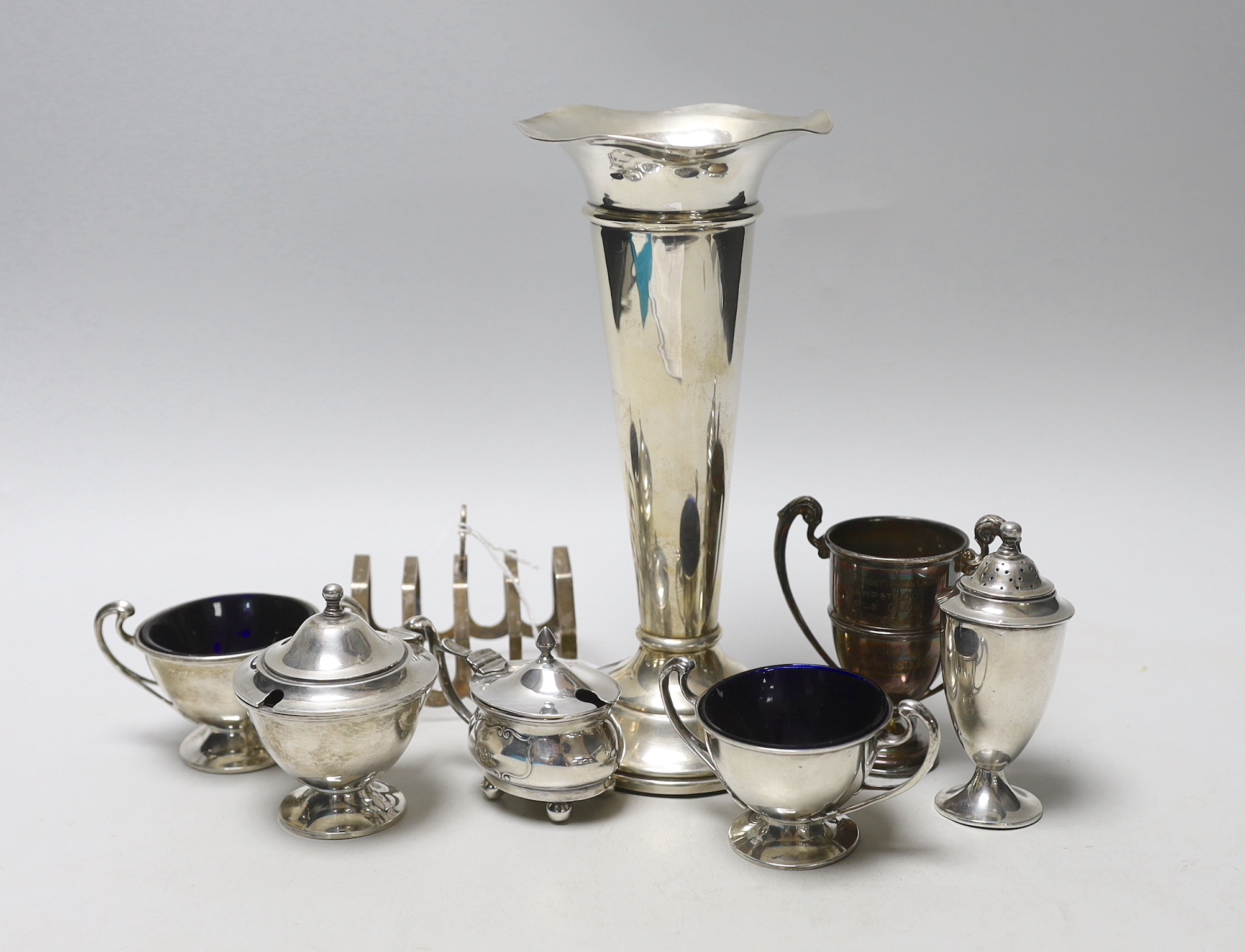 A George V silver three piece condiment set by Edward Barnard & Sons Ltd, London, 1916, two other silver condiments, silver toastrack, silver small trophy cut and silver spill vase.                                       