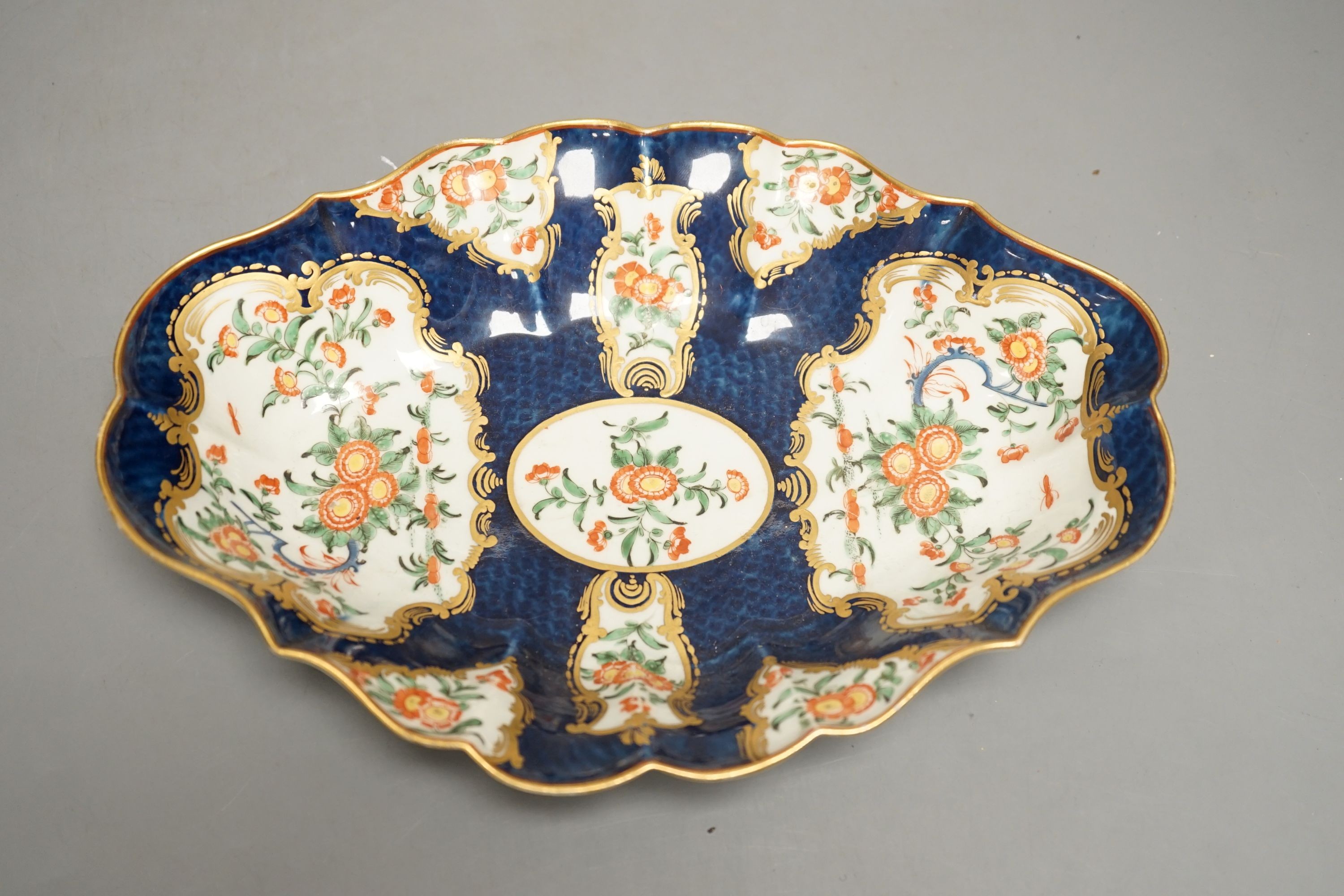 A Worcester scalloped oval dish with blue scale ground painted in kakiemon style with oriental flowers in git panels c.1768, 25 cms wide.                                                                                   