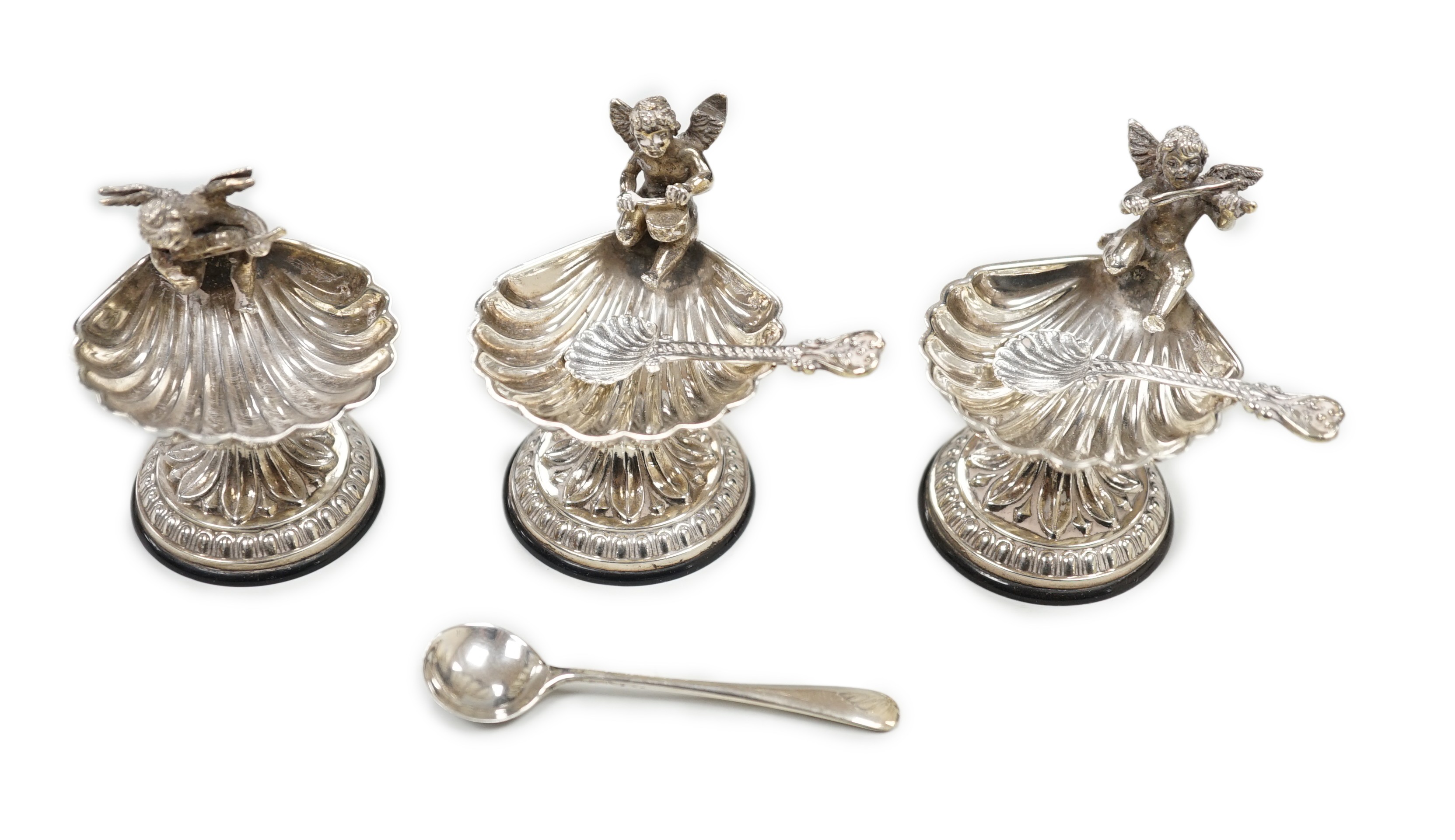 A set of three 20th century Spanish white metal shell pedestal salts with cherub handles and three spoons, height 50mm.                                                                                                     