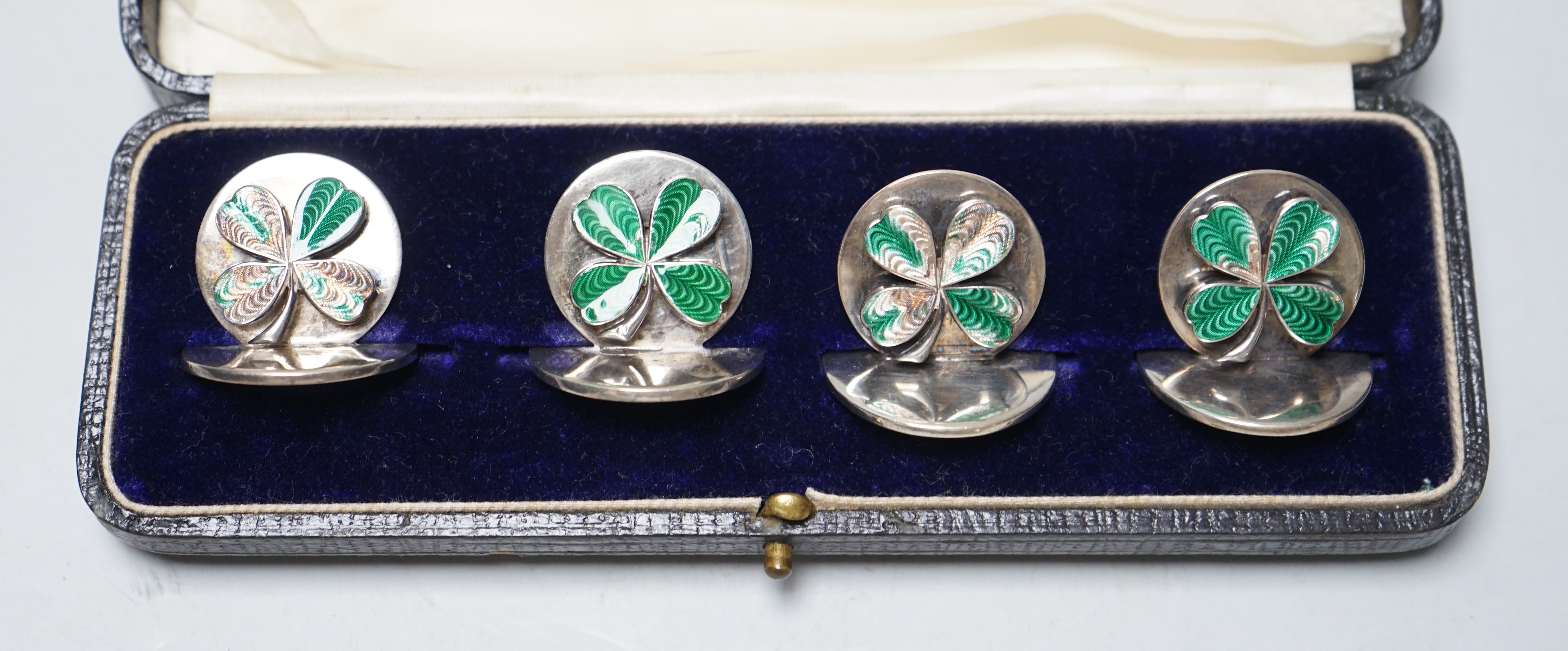 A cased set of four George V enamelled silver menu holders, decorated with four leaf clovers, by Sampson Morden & Company (a.f.), diameter 31mm.                                                                            