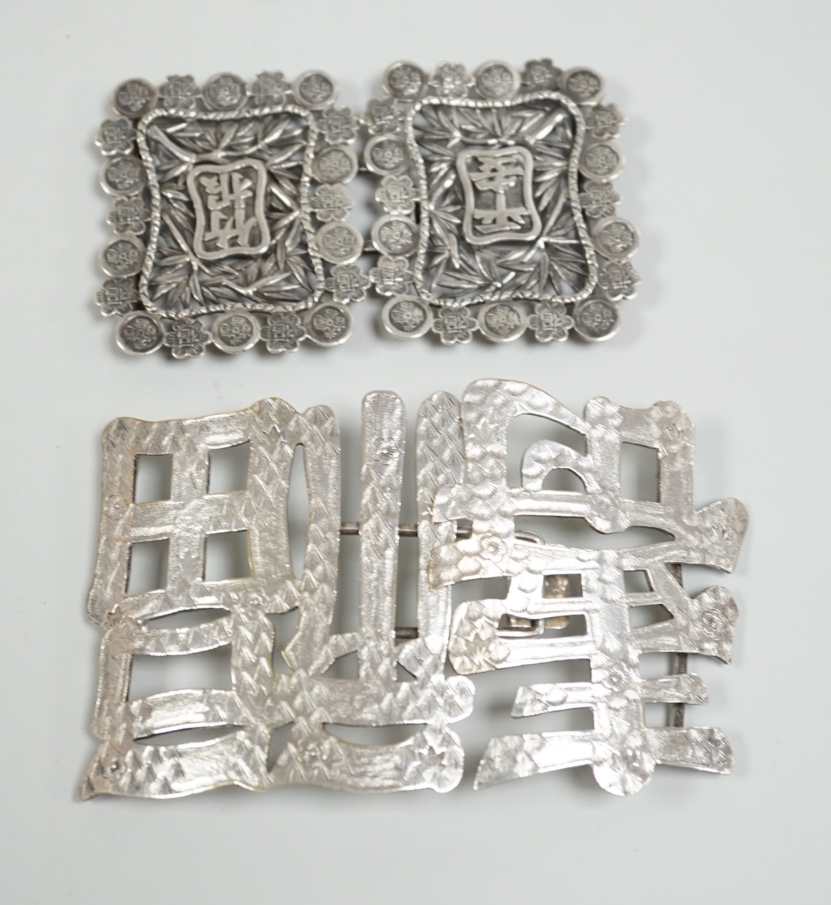 A Chinese white metal belt buckle, 13.3cm wide, the border cast with models of Guangxu cash and a Chinese plated ‘fu Shou’ belt buckle                                                                                      