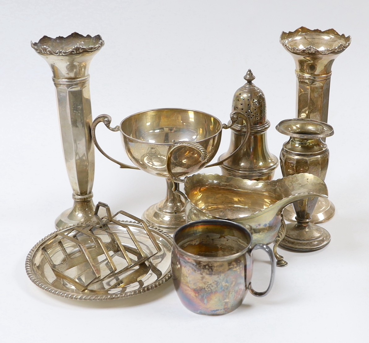 Assorted silver ware including a pair of George V spill vases, a small vase, a two handled trophy cup, sauceboat, mug, caster, stand and toastrack.                                                                         