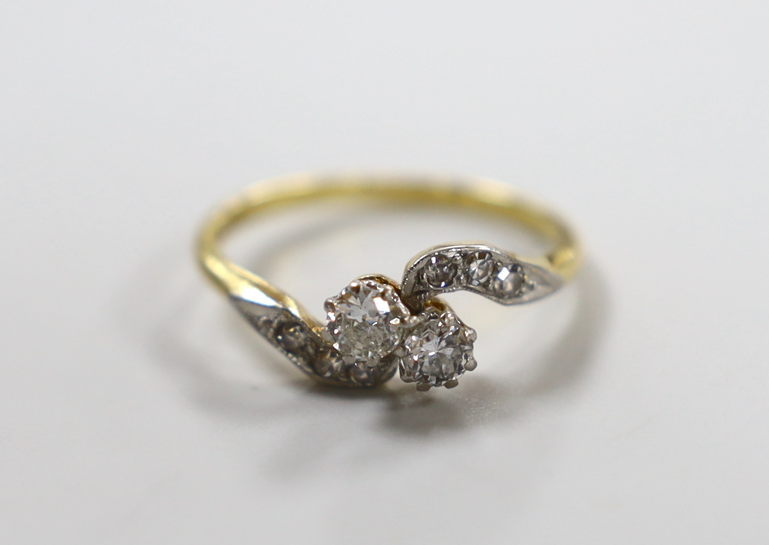 An 18ct, pt and two stone diamond set crossover ring, with diamond chip set shoulders, size P, gross weight 2.8 grams.                                                                                                      