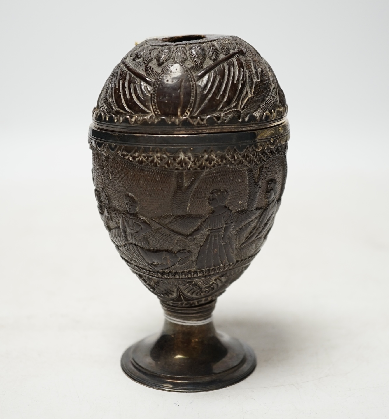 An early 19th century white metal mounted coconut cup, carved with continuous scene of figures, height 13.8cm. Condition - poor                                                                                             