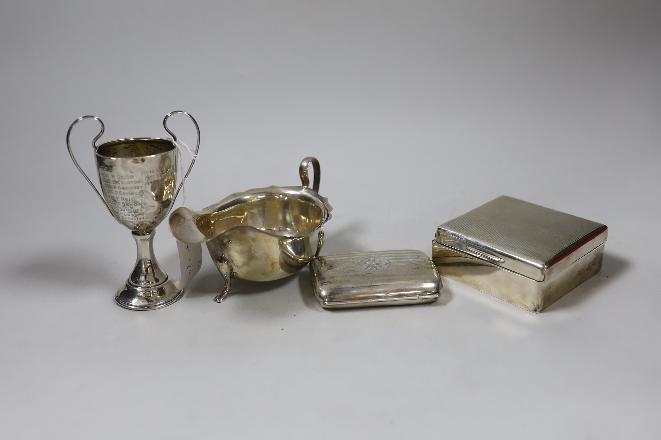 A George V silver sauceboat, a small silver cigarette box, a similar case and a small silver two handled trophy cup.                                                                                                        