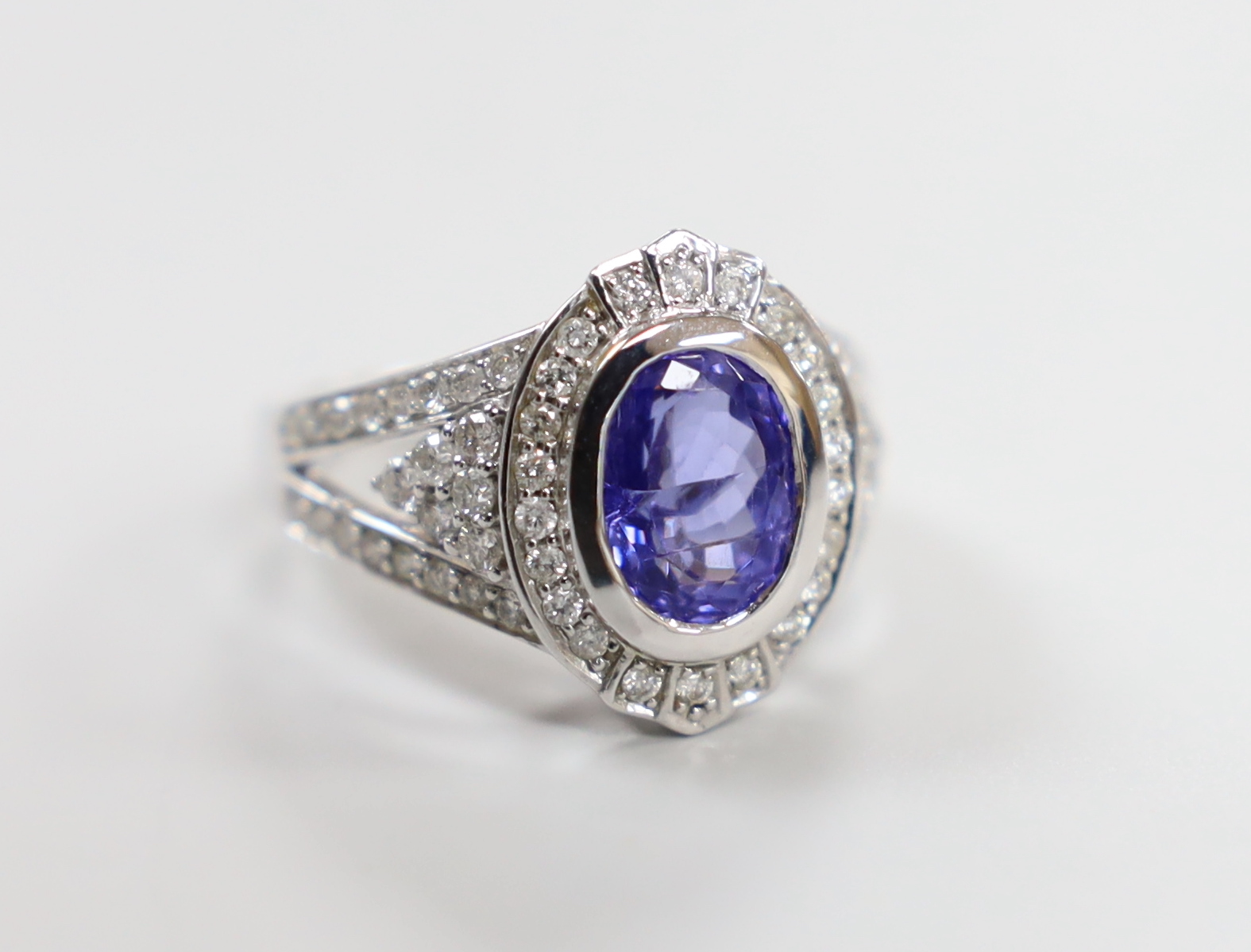 A modern 14k white metal, oval cut tanzanite and diamond chip set cluster dress ring, with diamond chip set shoulders, size M, gross weight 7 grams.                                                                        
