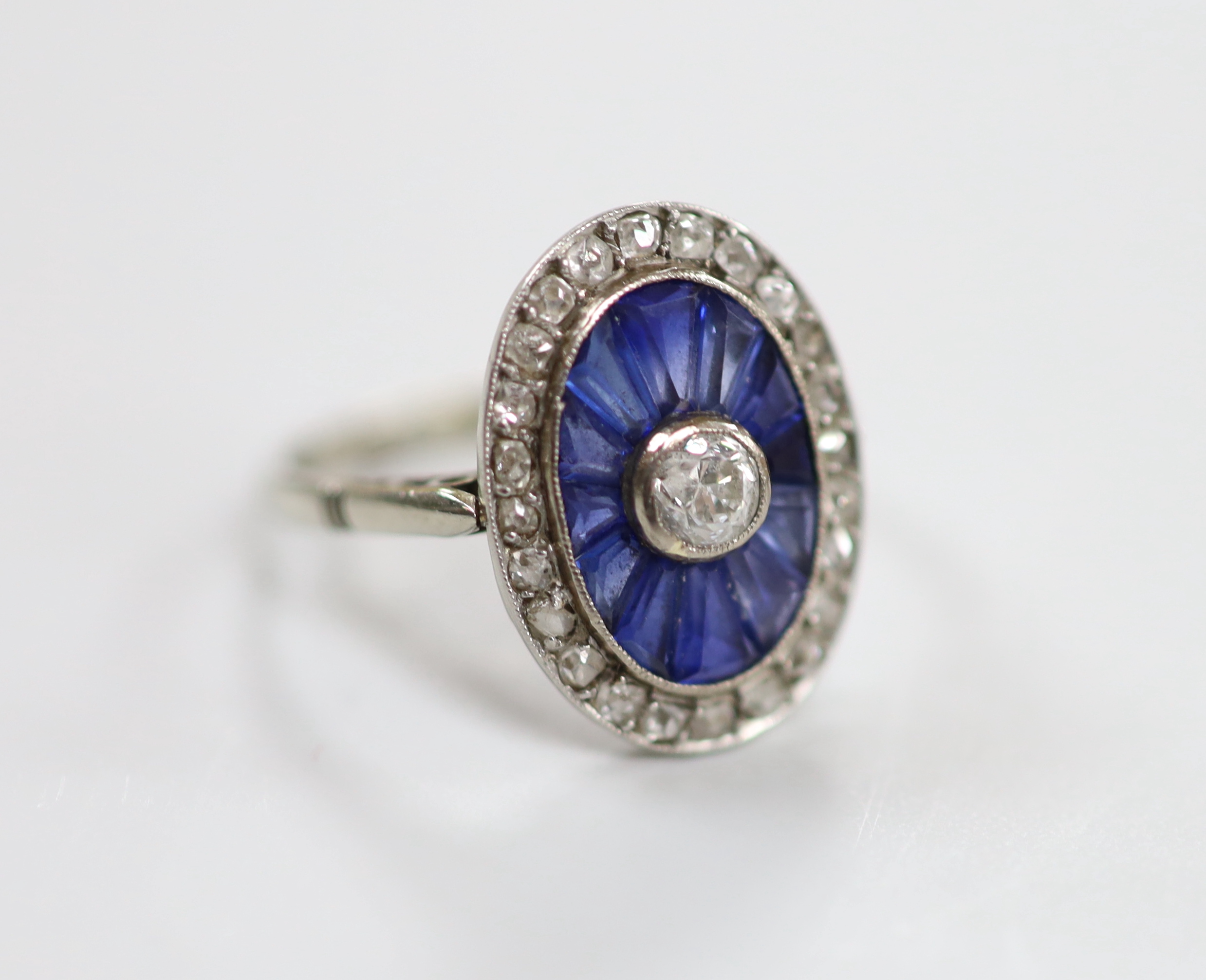 A 1920's white metal, sapphire and diamond oval cluster ring, set with round cut diamonds and shaped cut sapphires, size M/N, gross weight 3.3 grams.                                                                       