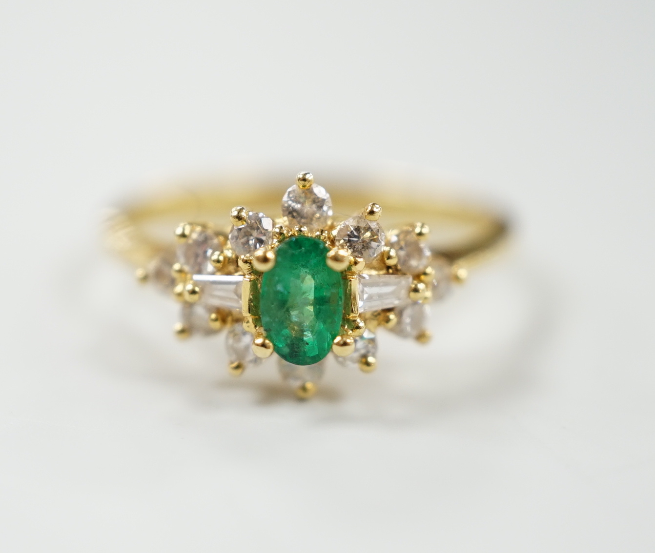 A modern 18ct gold, single stone oval cut emerald and round and trapeze cut diamond set cluster ring, size M, gross weight 2.8 grams.                                                                                       