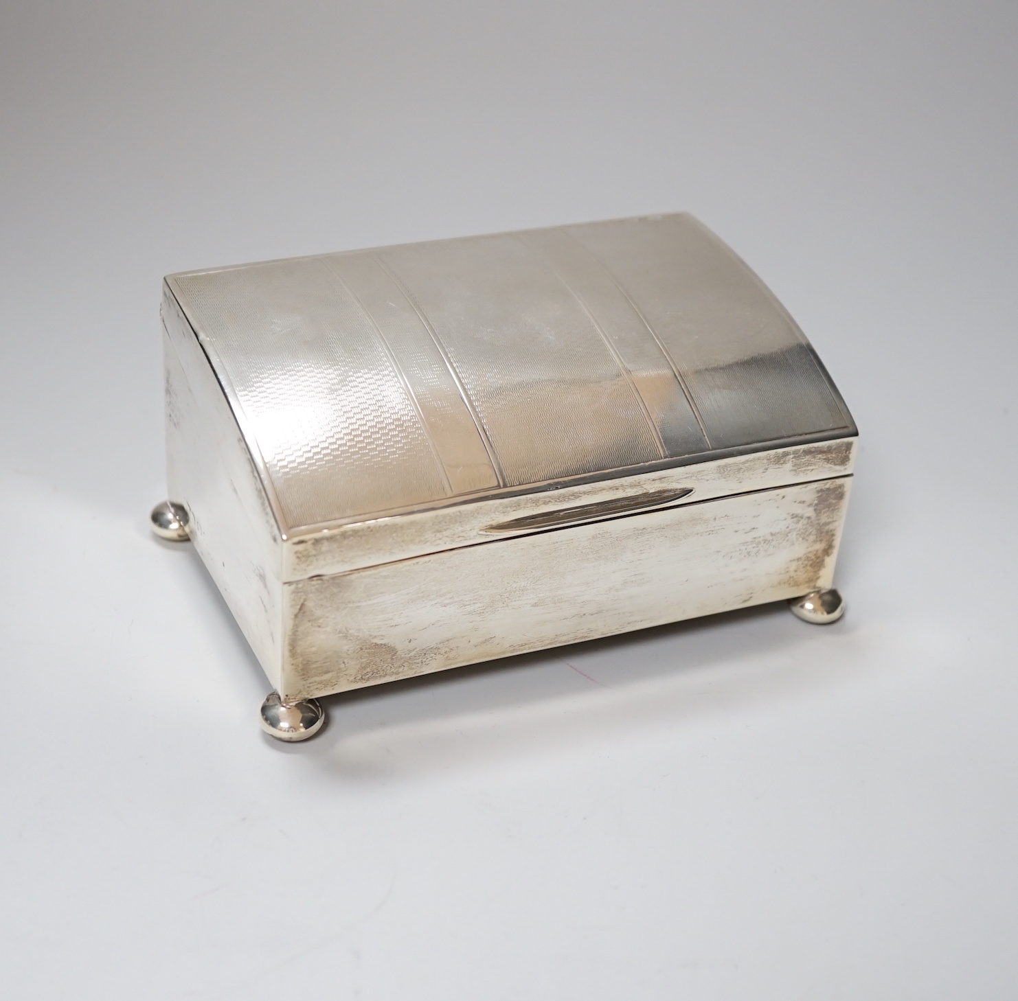 A George V engine turned silver cigarette box, of unusual sloping lidded form, with pad feet, marks rubbed, 14cm, gross 485 grams                                                                                           