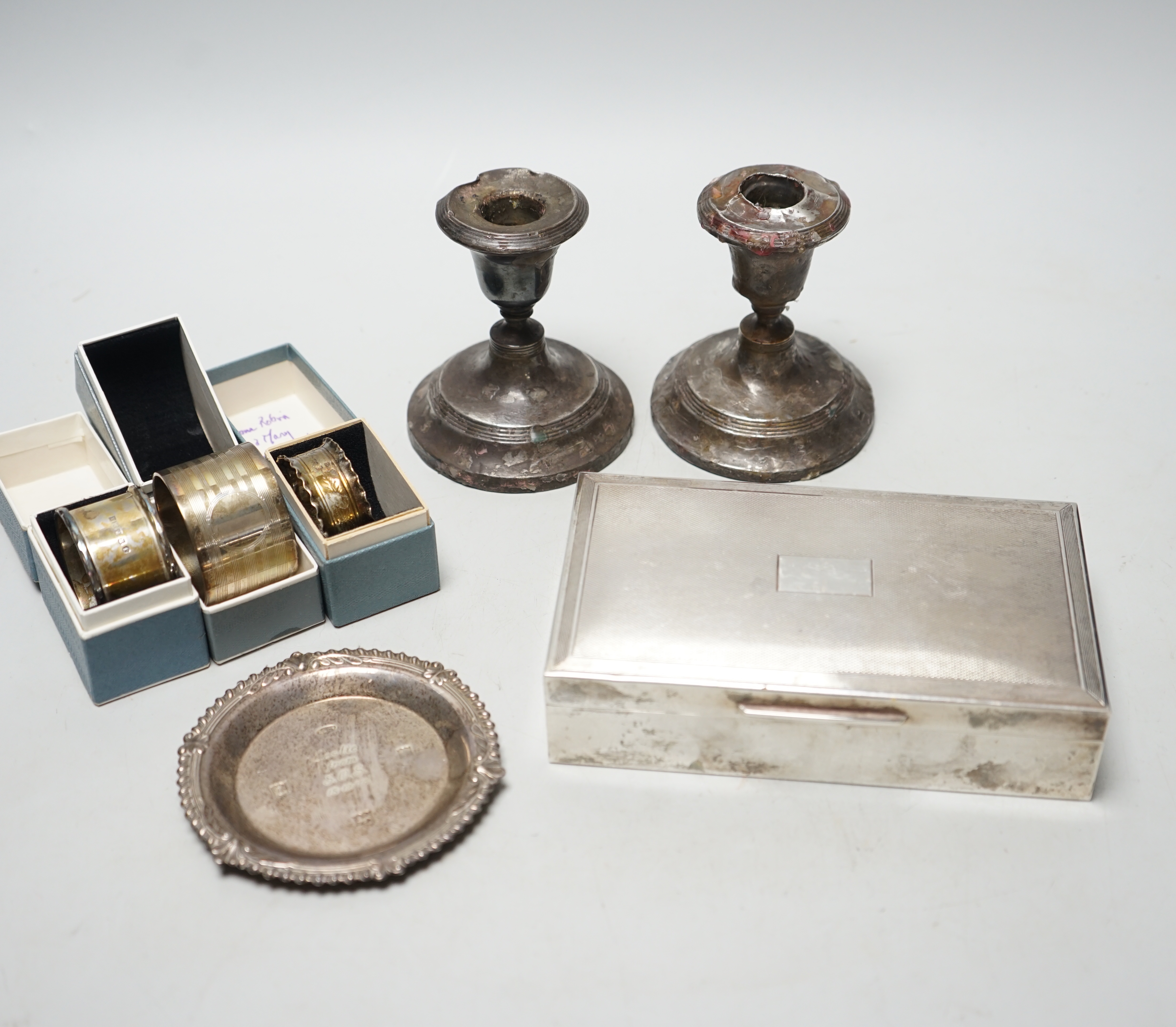 A 1960's engine turned silver mounted rectangular cigarette box, three boxed silver napkin rings, a small silver dish and a pair of damaged silver mounted dwarf candlesticks.                                              