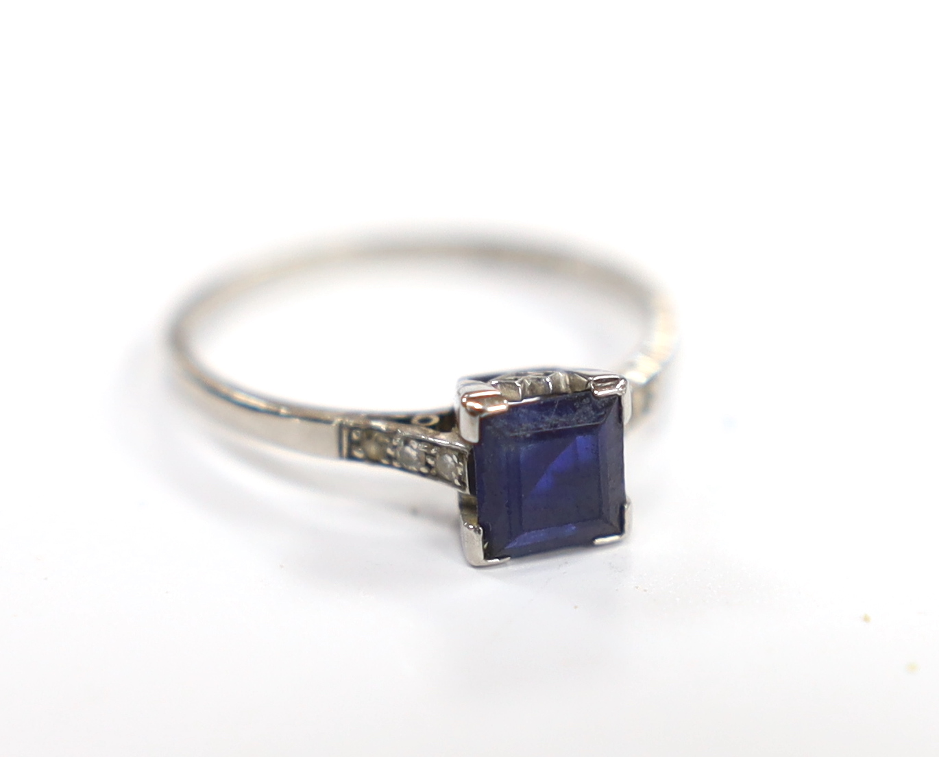 A white metal (stamped plat) and single stone square cut sapphire set ring, with diamond chip set shoulders, size P, gross weight 2.8 gram.                                                                                 