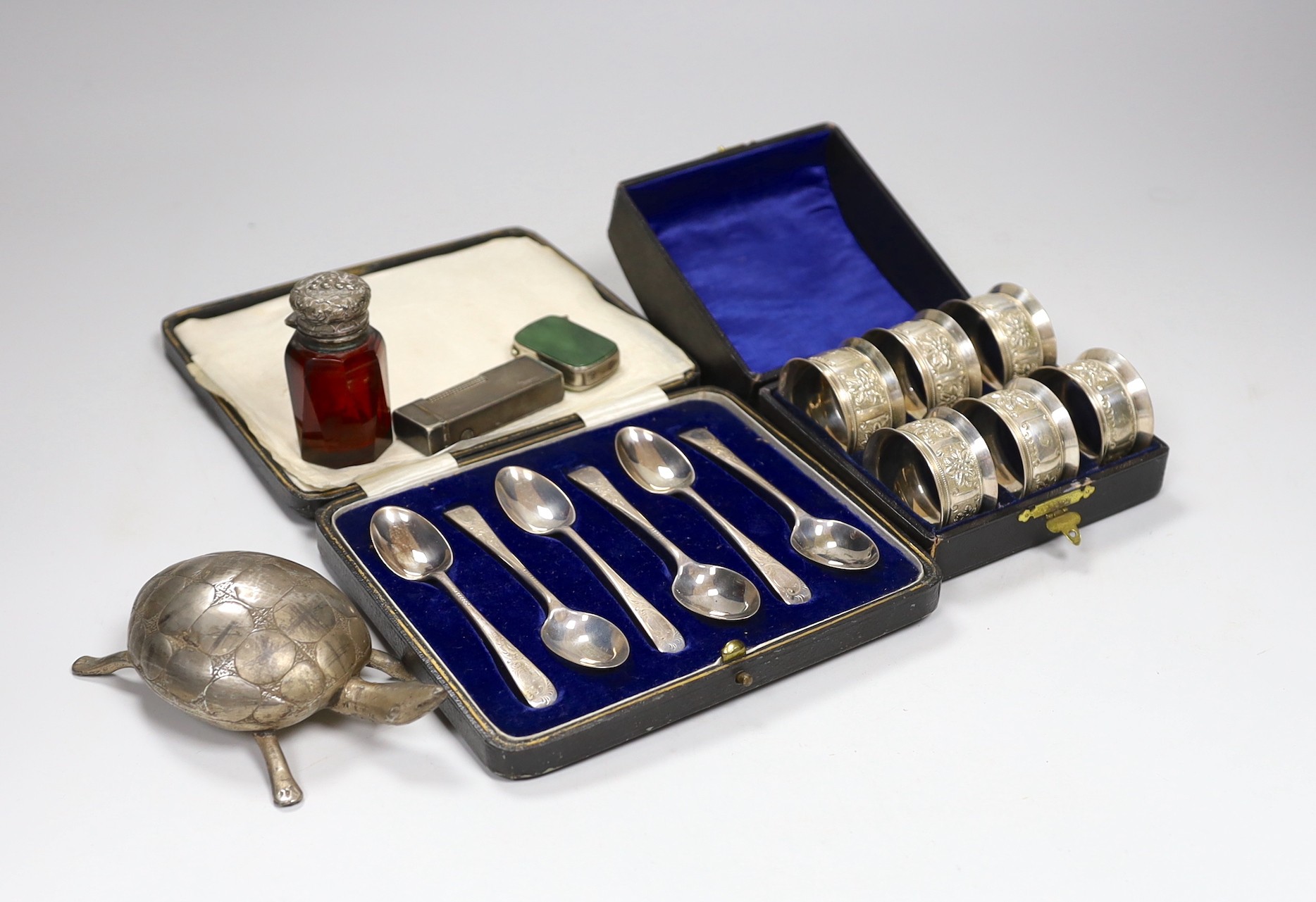 A cased set of six George V silver teaspoons, a white metal mounted glass scent bottle, a Dunhill lighter, a plated vesta, a white metal tortoise and a cased set of six plated napkin rings.                               