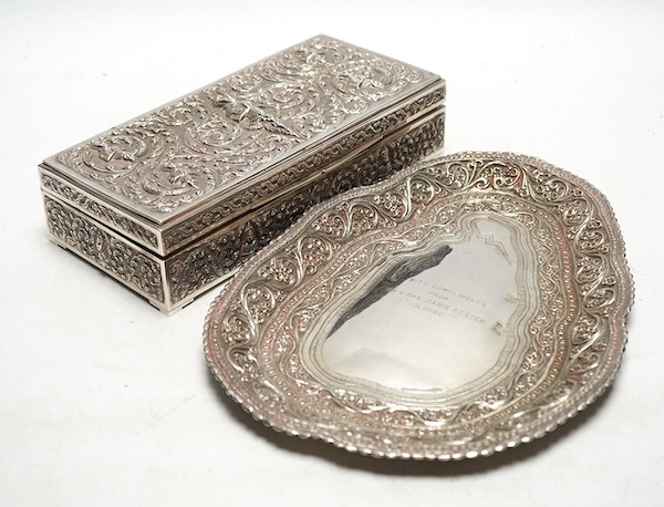 A Thai white metal cigarette box, wood lined, stamped Thainakon, 20cm and a Ceylonese white metal dish, shaped in the form of the country, with embossed borders, and inscription, maker’s mark SS, 24.7cm long, 182g. Condi